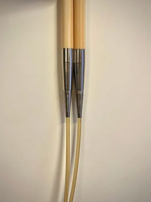 Close-up of two wooden knitting needles with metal connectors and yellow cables attached, aligned side by side vertically against a light background.