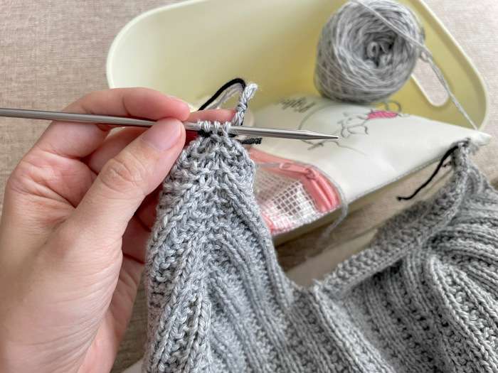 Close-up photo of gray yarn camisole being knitted, showing stitches on needles, yarn ball, and a knitting pouch in the background.